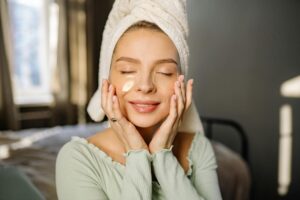 Mujer sonriente con una toalla en la cabeza y parches para los ojos, disfrutando de su rutina de autocuidado facial en casa.